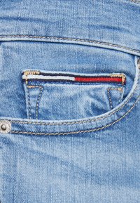 Jeans de mezclilla azul claro con un pequeño bolsillo decorativo que presenta rayas rojas, blancas y azules. Incluye un detalle de botón de latón y costuras en contraste.