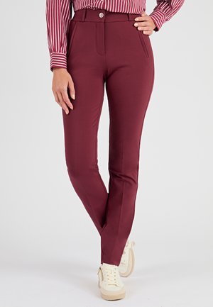 Pantalons ajustés bordeaux avec une texture lisse, dotés de poches latérales et d'une fermeture à bouton à l'avant, associés à des baskets crème.