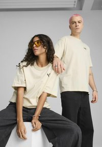 T-shirt oversize beige abbinate a pantaloni in denim neri. Le t-shirt presentano un piccolo logo. Un modello indossa grandi occhiali da sole gialli, l'altro ha i capelli rosa.