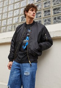 Sort bomberjakke med ribbet krave og manchetter, parret med en grafisk t-shirt og slidte blå jeans. Guld lynlåsdetaljer er synlige.