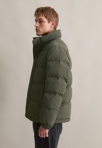Jachetă puffer verde măsliniu cu guler înalt, având secțiuni quilted și manșete elasticizate. Finitură subtilă și design ușor.