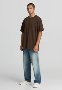 T-shirt in cotone marrone con vestibilità rilassata e maniche corte, abbinato a jeans in denim blu medio e sneakers bianche. Design minimalista, senza fantasie.