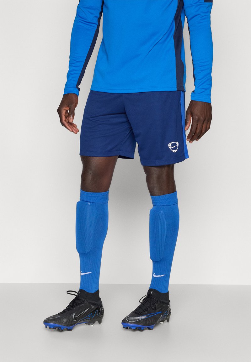 Μπλε αθλητικό πουκάμισο με μακριά μανίκια και ναυτικές λεπτομέρειες, συνδυασμένο με ναυτικά σορτς, μπλε κάλτσες μέχρι το γόνατο και μαύρα παπούτσια με μπλε λεπτομέρειες.