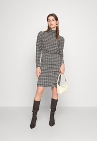 Rochie în carouri alb-negru, cu mâneci lungi, gât înalt, talie strânsă, asortată cu cizme maro până la genunchi și o poșetă mică de crem.