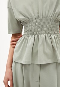 Helles grünes Oberteil mit kurzen Ärmeln, tailliertem Schnitt und ausgestelltem Peplum-Saum. Hergestellt aus weichem Stoff mit dezenten Raffungselementen.