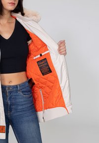 Hellblaue Bomberjacke mit einer orangefarbenen, gesteppten Fütterung, fellbesetzter Kapuze und Frontreißverschluss. Verfügt über ein Etikett und seitliche Taschen.