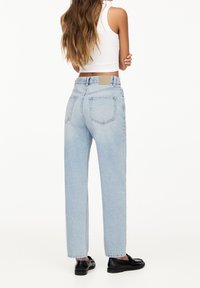 Donna con lunghi capelli che indossa jeans high-waisted blu chiaro, top bianco senza maniche e mocassini neri, in piedi con le braccia incrociate, voltata di spalle.