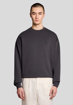 Jonge persoon met kort haar die een donkergrijze lange mouwen trui en cremewit geplooide broek draagt tegen een effen achtergrond.