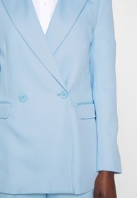 Blazer doppiopetto azzurro chiaro realizzato in tessuto testurizzato. Presenta un colletto a punta, due bottoni blu e due tasche laterali.