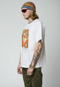 T-shirt blanc à manches courtes avec un imprimé graphique coloré présentant du texte orange et vert, porté avec un pantalon vert olive et un bonnet en tricot multicolore.