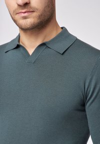 Strick-Polohemd in dunklem Teal, mit V-Ausschnitt und kurzem, strukturiertem Kragen. Glatte Textur mit geripptem Detail am Kragen.