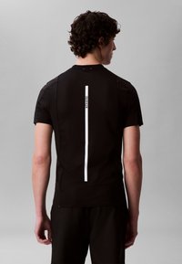 Mann sett bakfra, iført en svart kortermet skjorte med en vertikal hvit stripe og liten "Calvin Klein"-tekst på ryggen.