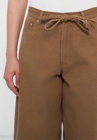 Brune vide bukser lavet af tekstureret stof, med en bundet talje, forlommer og synlige syninger.