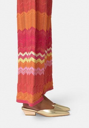 Piernas inferiores con una falda larga de patrón zigzag multicolor y mules doradas con tacón bajo y punta puntiaguda sobre un fondo liso.