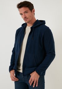 Felpa con zip blu navy dal tessuto morbido, con tasche frontali e lacci sul cappuccio, indossata sopra una t-shirt chiara e jeans.