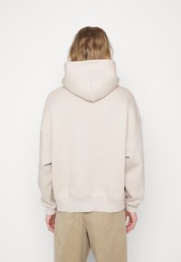 Personne aux cheveux longs portant un sweat à capuche beige clair et un pantalon beige, debout de dos sur un fond blanc uni.