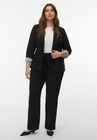 Zwarte blazer en bijpassende broek met een ceintuur in de taille, gecombineerd met een wit topje. Gestructureerde manchetten en puntige zwarte schoenen maken de look compleet.