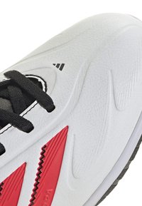Zapatilla deportiva blanca con parte superior de sintetico texturizado, cordones negros y acentos rojos; presenta un logo prominente y líneas de diseño elegantes.