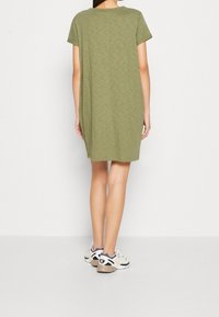 Robe vert olive à manches courtes avec une coupe décontractée, présentant une couture centrale au dos. Portée avec des chaussures de sport de couleur claire.