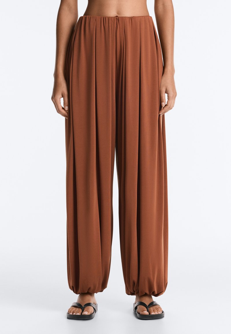 Pantalon large marron avec ourlets froncés porté par une personne debout, assorti à des sandales noires à bout ouvert sur fond blanc.