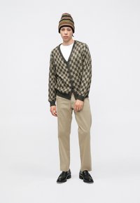 Cardigan a quadretti marroni e beige, tessuto lavorato a maglia, indossato sopra una t-shirt bianca. Pantaloni beige e scarpe nere completano il look.