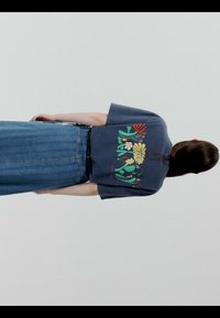 Personne aux longs cheveux foncés portant un T-shirt bleu foncé avec un motif floral coloré et une jupe en jean taille haute, vue de dos.