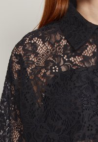 Svart bluse i blonde med blomstermønster, delikat tekstur og klassisk krage. Stoffet er gjennomskinnelig med intrikate detaljer.