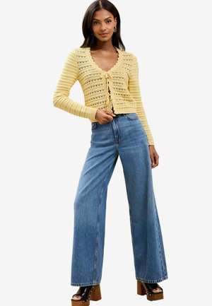 Gelbes gestricktes Cardigan mit einem gewellten Halsausschnitt, kombiniert mit hoch-taillierten, weit geschnittenen blauen Jeans und braunen Plateaupumps.