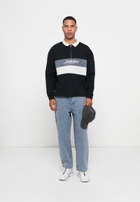 Zwarte pullover met een witte en grijze horizontale streep, ritskraag en logo. Draag het met lichtblauwe jeans en witte sneakers. Houdt een grijze pet vast.