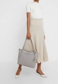 Grijze leren tote bag met dubbele handvatten, ritssluiting, logo-accent en een gladde textuur, gecombineerd met een beige ribgebreide rok.