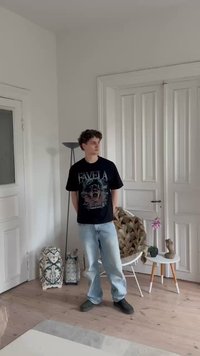 Melnā T-kreklā ar "FAVELA" grafiku, gaiši zili džinsi un melnas kurpes. Istabā ir rakstainas somas, lampa, augs un skulptūra.