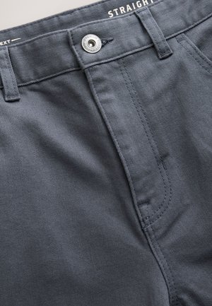 Tmavosivé nohavice so strečovým strihom z textúrovanej látky. Majú kovové gombíky a prešívanie pozdĺž pásu a oblastí so zipsom.