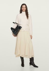 Haufige cremefarbene Strickjacke mit V-Ausschnitt, kombiniert mit einem fließenden cremefarbenen Rock. Schwarze Lederhandtasche mit einzigartiger Form und Textur. Knielange schwarze Stiefel.