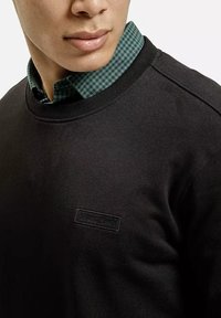 Czarna bluza z okrągłym dekoltem wykonana z bawełny o teksturowanym wykończeniu. Zielona koszula w kratę z kołnierzykiem wystaje spod spodu.