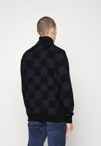 Maglione nero con colletto alto presenta un motivo a scacchi. Realizzato in un tessuto testurizzato, ha una vestibilità aderente e un elastico in vita.