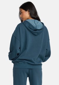 Oversized hoodie in dark teal, featuring a drawstring hood and soft texture. The interior hood is lighter blue, with ribbed cuffs and hem.