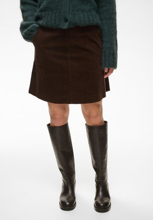 Personne portant une jupe en velours côtelé marron, un pull en maille vert foncé et des bottes en cuir noires montant jusqu'aux genoux, debout sur un fond blanc.