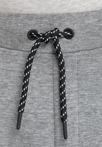 Lichte grijze joggers met een zwarte koordsluiting met witte accenten, een geribbelde textuur en een elastische tailleband. Close-up van de koordsluiting.