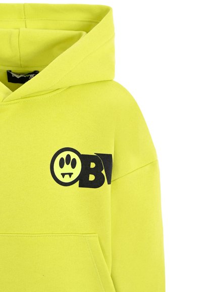 Heldere gele hoodie van zacht materiaal, met een voorzak en een zwarte grafische afdruk van een lachend gezicht en het "OBV"-logo.