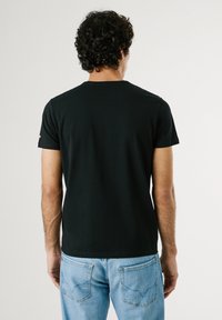 Homme aux cheveux bouclés portant un t-shirt noir uni à manches courtes et un jean bleu clair, debout de dos sur un fond clair.