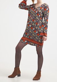 Robe longue à manches longues à fleurs, avec un col en V, présentant un fond sombre, des fleurs orange et blanches, et un ourlet à motifs. Bottines marron.
