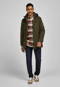 Olijfgroene waterdichte jas met capuchon, voorkeursrits en twee zijzakken, gedragen over een rood en grijs geruit shirt, gecombineerd met donkere jeans en sneakers.