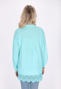 Camisa de manga larga azul claro con espalda plisada y dobladillo de encaje con forma de concha. Tela suave, corte relajado, combinada con jeans de denim claros.