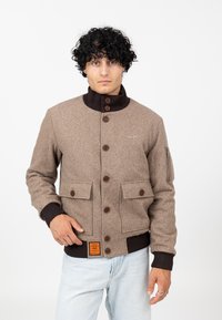 Mann mit lockigem schwarzen Haar, der eine braune Bomberjacke mit Knöpfen und großen Fronttaschen sowie eine hellblaue Jeans trägt, vor einem weißen Hintergrund.