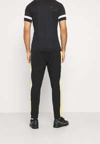 Schwarzes sportliches Shirt und Hose mit gelben seitlichen Streifen. Das Shirt hat einen Rundhalsausschnitt, während die Hose über einen elastischen Bund und Bündchen verfügt.