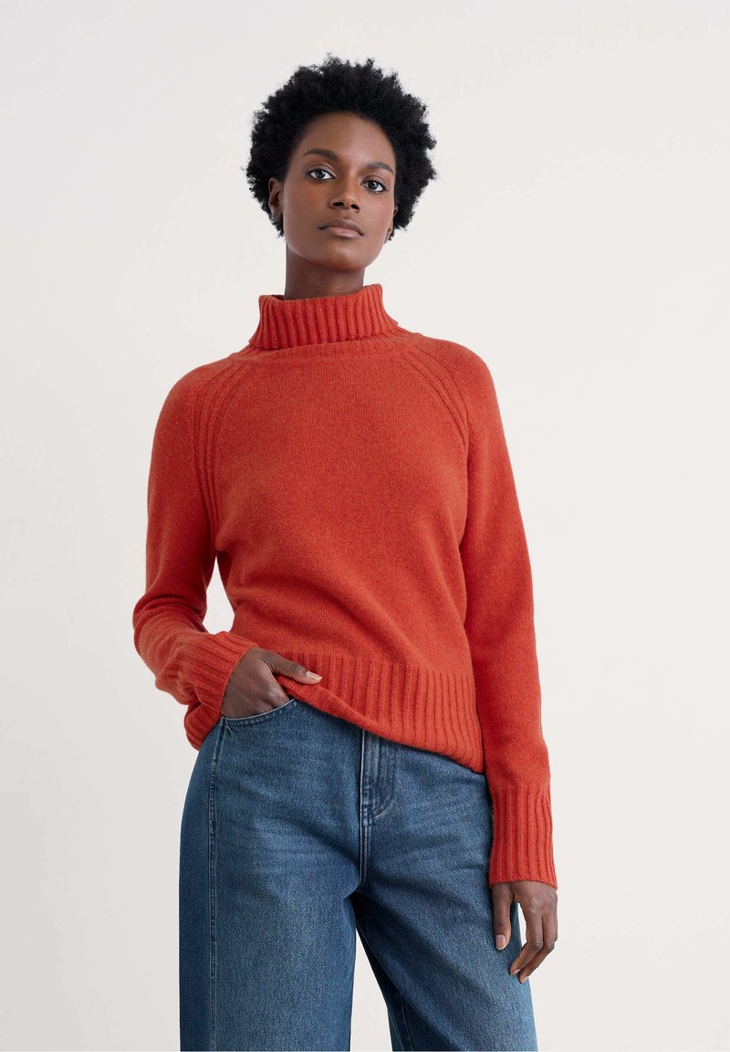 Femme portant un pull à col roulé couleur rouille et un jean bleu, avec une main dans sa poche, devant un fond clair uni.