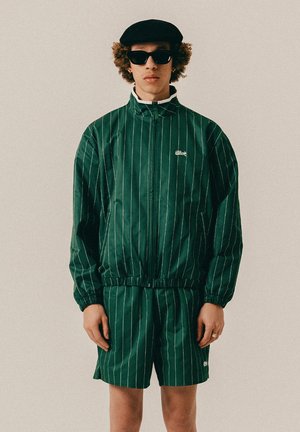 Ung person iført en mørkegrøn jakkesætjakke med nålestriber og shorts, sort baret og solbriller, stående mod en ensfarvet baggrund.