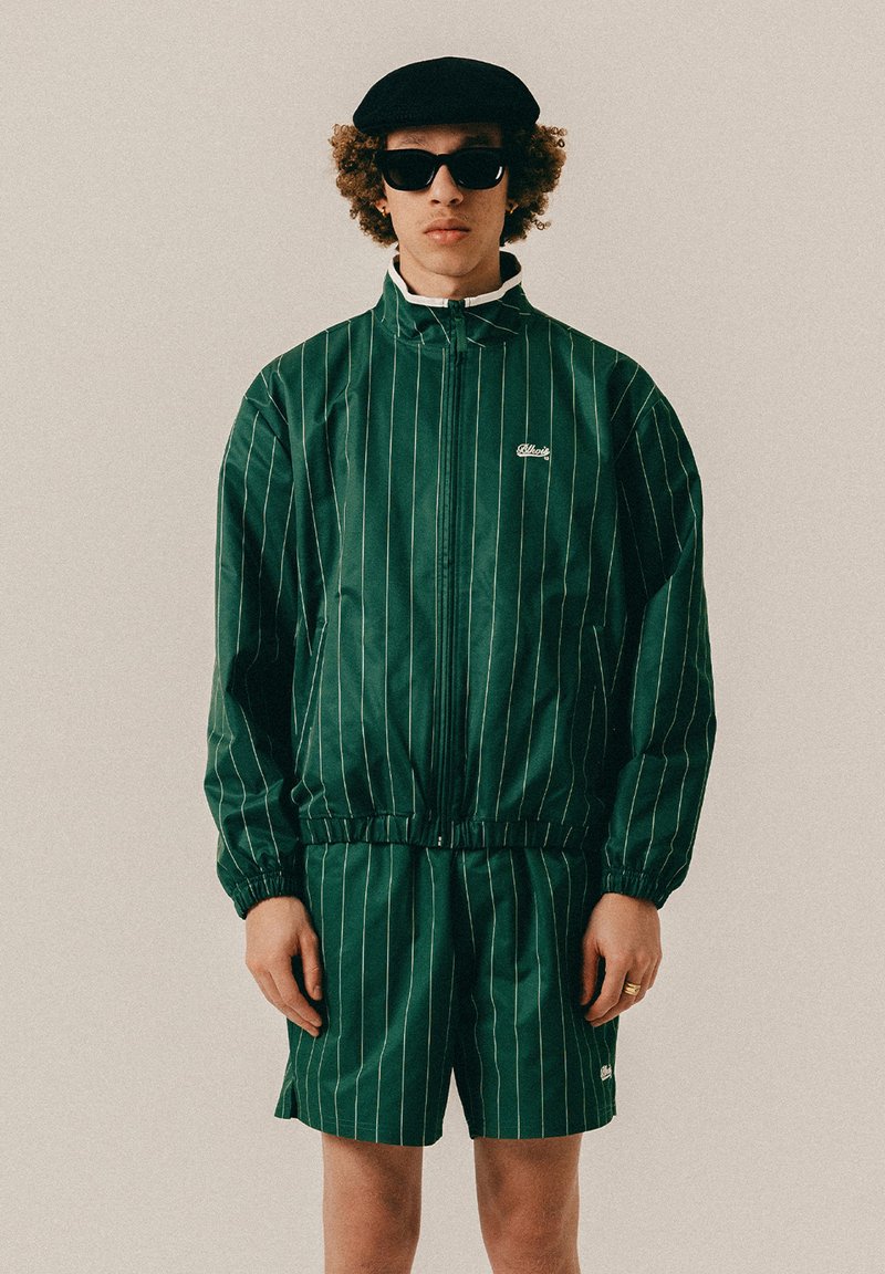 Młoda osoba ubrana w ciemnozieloną, prążkowaną kurtkę dresową i spodenki, czarny beret oraz okulary przeciwsłoneczne, stojąca na jednolitym tle.