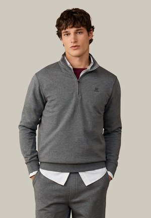 Jonge man met krullend bruin haar die een grijze halve rits trui draagt over een wit overhemd en bijpassende grijze broek, staand tegen een effen achtergrond.