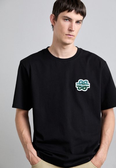 Black cotton t-shirt featuring a green and white embroidered logo on the left chest. Round neckline and short sleeves.